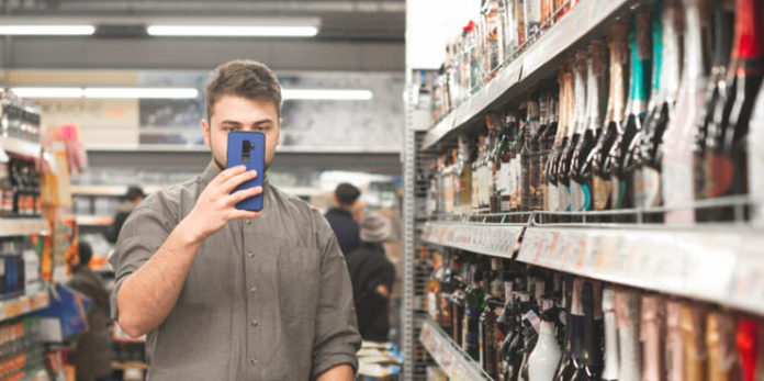 Man in a shirt stands in a supermarket with a shopping cart and holds a smartphone in his hands. Buyer with a smartphone makes a purchase at a supermarket, is in the department of strong drinks